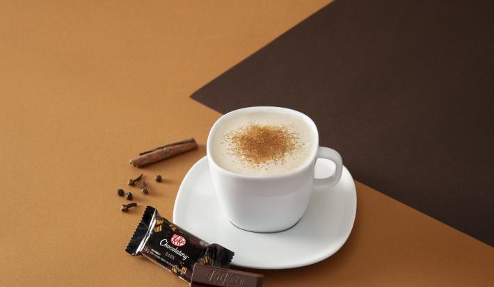 Foto da receita de cappuccino chai cremoso servida em uma xícara branca com um kit kat dark ao lado em fundo marrom claro e escuro