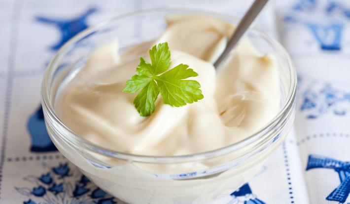 Imagem de um pequeno recipiente redondo de vidro contendo um creme branco. Uma folha de salsinha decora o centro do creme. O Recipiente está disposto sobre uma toalha em tons de branco e azul