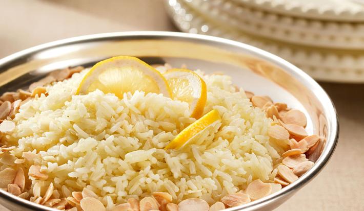 Fotografia em tons de prata e bege, com recipiente de inox com arroz cozido, amêndoas e limão-siciliano, ao fundo pratos empilhados com talheres por cima de um guardanapo, decoração com vela, um copo, tudo sobre bancada de cor bege.