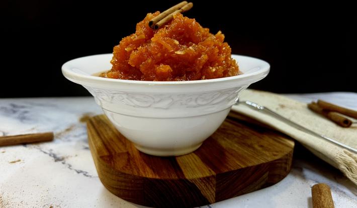 Foto da receita do doce de abóbora com coco servida em um bowl de porcelana branco e com um pau de canela decorativo em cima. O bowl está disposto em cima de uma tábua de madeira, esta em cima de uma mesa de mármore com mais paus de canela decorativos.