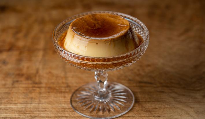 Fotografia de um mini flan de cappuccino dentro de uma decoração de vidro alta, sobre uma mesa em tons de madeira.