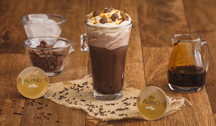 Fotografia em tons de marrom em uma bancada de madeira com um copo de vidro grande e a bebida de chocolate Alpino dentro. Ao fundo, um pote com chantilly, outro com a calda de caramelo e outro com gotas de chocolate e as cápsulas Dolce Gusto Alpino.