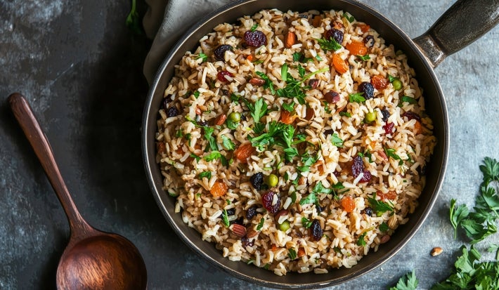 Em cima de uma mesa, frigideira com arroz soltinho uvas-passas pretas e brancas, decorado com folhas de cheiro-verde.