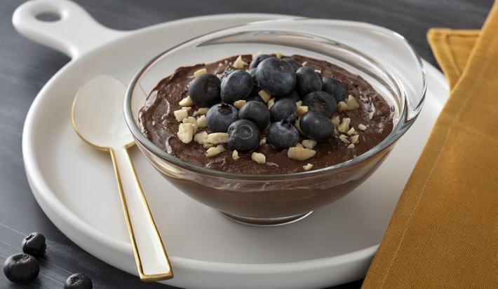 Foto em tons escuros da receita de mousse de chocolate com mirtilo servida em uma taça de vidro sobre uma frigideira de porcelana branca com uma colher branca ao lado