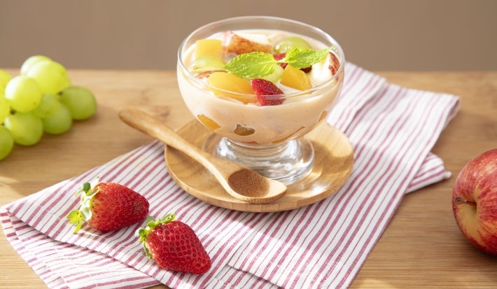 Fotografia em tons claros de um recipiente pequeno decorativo com as frutas e o creme sobre um apoio circular de madeira pequeno ao lado de uma colher pequena de madeira.