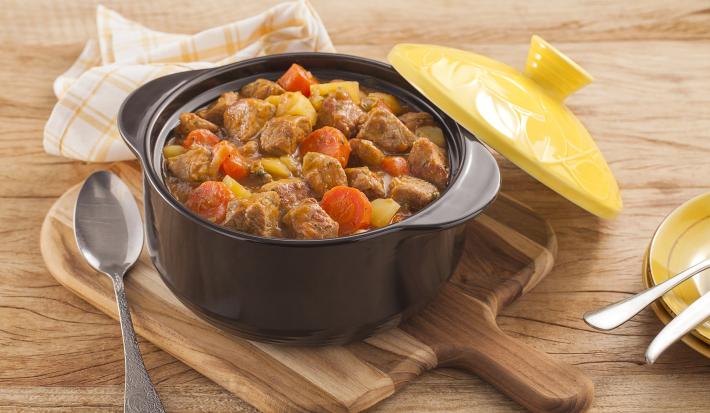 Fotografia em tons de amarelo em uma bancada de madeira clara, um pano amarelo com listras, uma tábua de madeira, uma panela preta de ferro com o picadinho de carne suína com legumes dentro e uma tampa amarela ao lado.