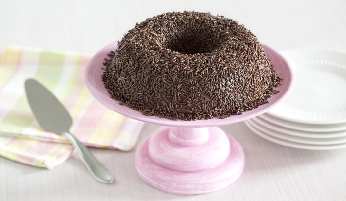 Foto de um brigadeirão de chocolate em cima de um prato de bolo rosa e com pratos brancos atrás