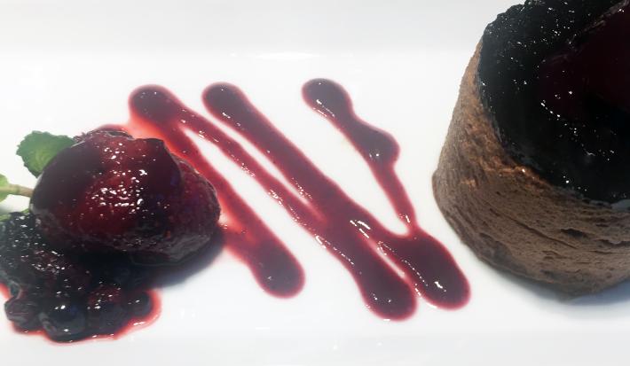 Fotografia de um flan de chocolate sobre um prato branco com uma calda de frutas vermelhas e pequenas folhas de hortelãs.