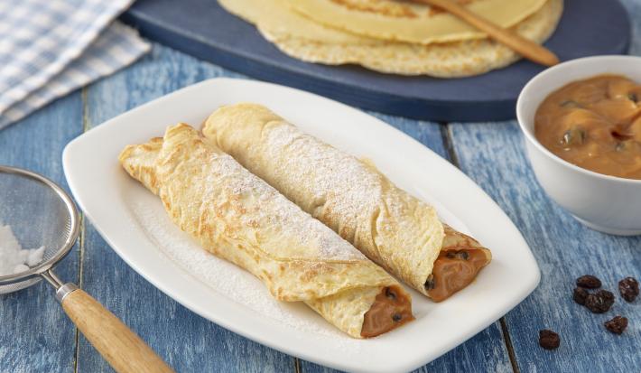 Foto da Receita de Panqueca de Doce de Leite. Observa-se um prato quadrado branco com duas panquecas em cima polvilhadas com açúcar. Do lado esquerdo, tem-se uma peneira com o açúcar e, do direito, as massas de panqueca inteiras.
