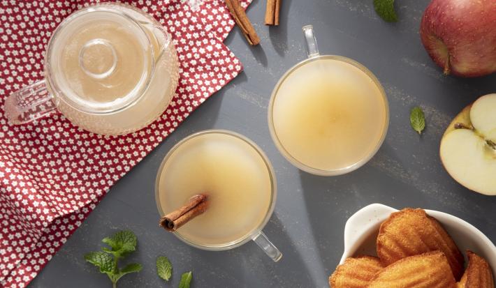 Imagem vista de cima de duas xícaras de chá de maçã, decorado com um pedaço de canela, maçãs e um pote de madeleines
