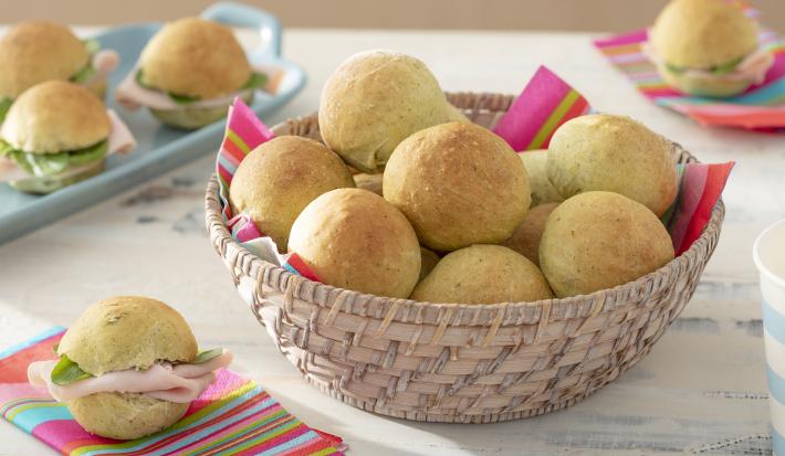 Foto da receita de Pãozinho de Talos. Observa-se uma cestinha com os mini pães dentro e, atrás, como decoração, alguns pães recheados de frios.