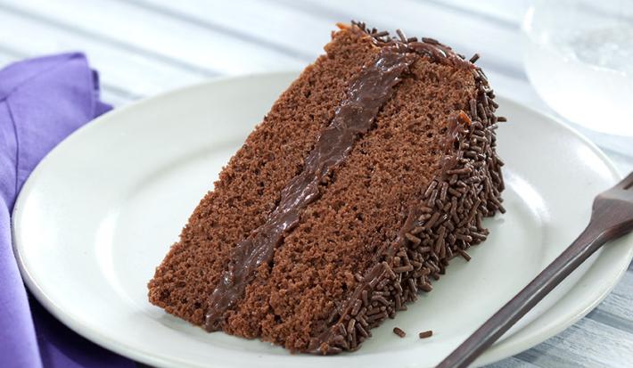 Foto de um prato branco numa mesa de madeira branca com uma fatia de bolo de chocolate dentro e um garfo do lado
