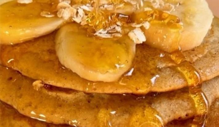 Fotografia cinco panquecas de baunilha empilhadas com mel, banana em rodelas e aveia por cima. As panquecas estão sobre um prato de vidro branco.