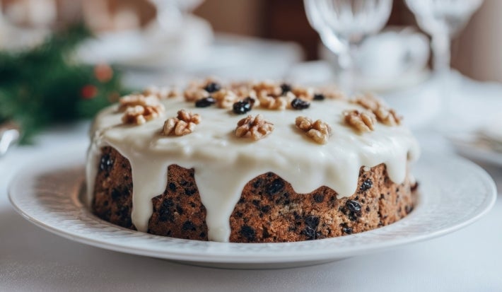 Fotografia de um bolo de frutas decorado com cobertura branca e nozes, apresentado em um prato elegante. O fundo sugere um ambiente festivo e natalino.