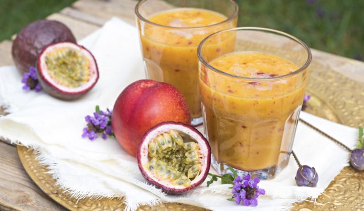 Fotografia de dois copos de vidro com uma bebida com suco de maracujá, leite e neston, apoiados sobre um pano fino branco, e por baixo, um sousplat. Ao lado dos copos, tem dois maracujás inteiros e uma metade aberta.
