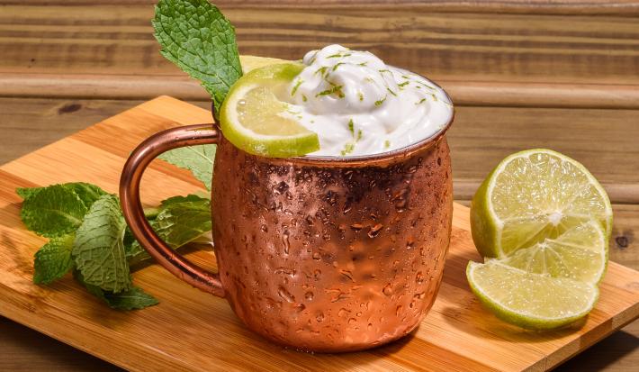 Foto da receita de Moscow Mule Sem Álcool. Observa-se uma caneca de alumínio rose gold com a bebida dentro e, por cima, a espuma. Limões e hortelã decoram a foto.