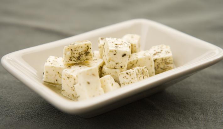 Fotografia de cubos de queijo branco fresco temperados e com azeite dentro de um recipiente pequeno, fundo e quadrado de vidro na cor branco. O aperitivo está sobre uma toalha de mesa preta.