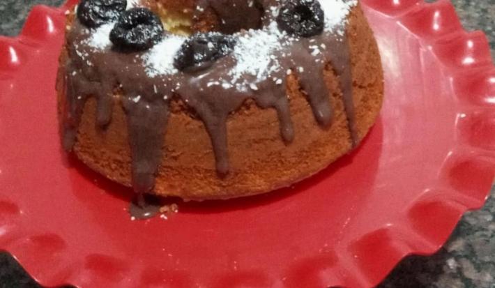 Fotografia de um bolo simples feito na forma de furo central, sobre um apoio grande vermelho decorativo. O bolo está com uma calda de brigadeiro, ameixas e coco ralado por cima.