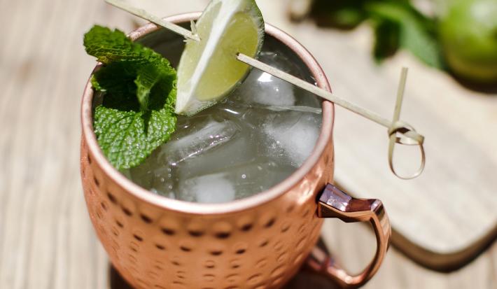 Foto da receita de Moscow Mule Blue, servida em uma caneca de estanho com gelo, sobre uma bancada de madeira decorada com algumas fatias de limão