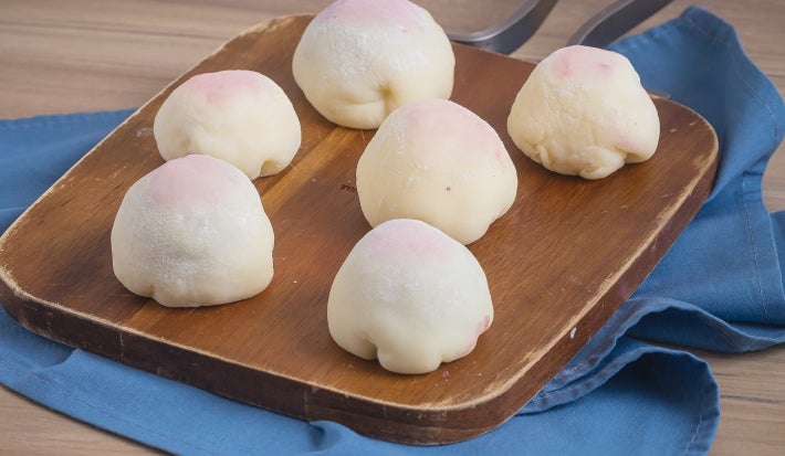Receita de Mochi de Brigadeiro, servida em pequenas bolinhas brancas, recheadas com brigadeiro enrolado, sobre uma tábua de madeira e um tecido na cor azul ao lado de uma Lata de Leite Moça