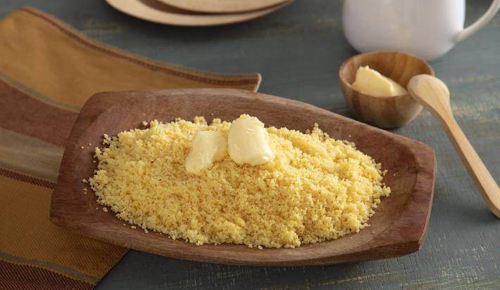 Foto da receita de Cuscuz de Milho. Observa-se uma tigela de madeira com o cuscuz soltinho e com manteiga em cima derretendo.