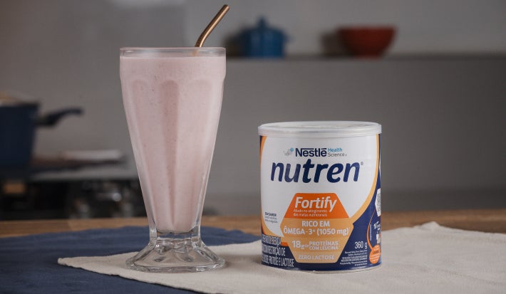 A imagem mostra o frozen de morango servido em um copo de vidro alto estilo de milkshake, com uma lata de nutren fortify ao lado, tudo sobre um pano bege em cima de uma mesa de madeira