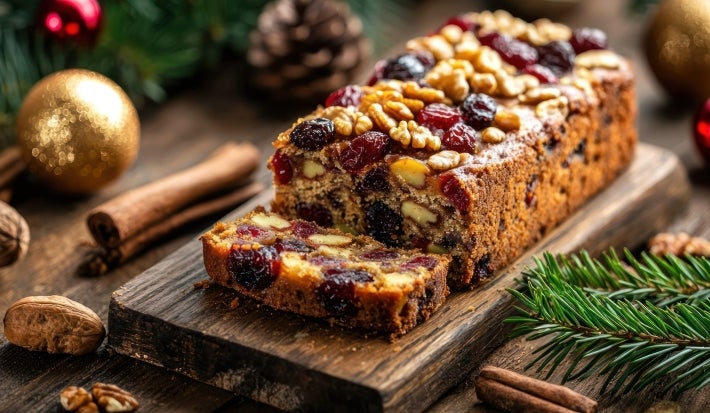 Fotografia de um bolo de frutas festivo, exibido em uma tábua de madeira rústica, repleto de frutas secas e nozes, cercado por elementos natalinos como ramos de pinheiro e canela.