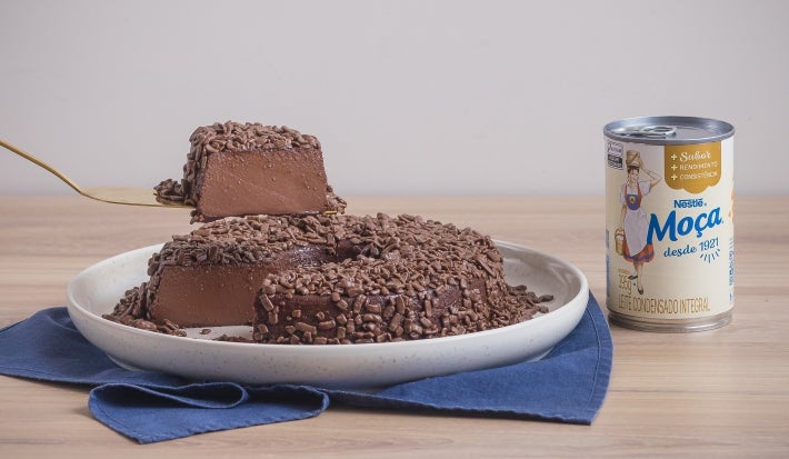 Foto da receita de Brigadeirão Moça, servido em um prato branco, decorado com muitos granulados de chocolate. O prato está entre uma lata de Leite Moça e uma espátula dourada, tudo sobre uma bancada de madeira