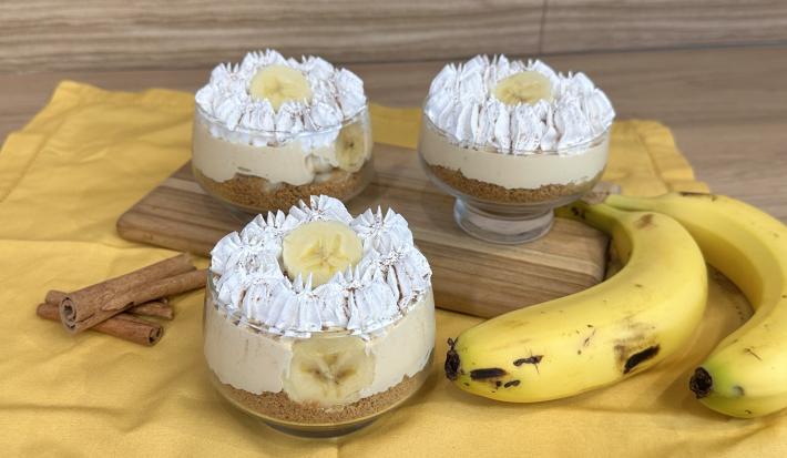 Fotografia em tons de amarelo com um pano amarelo ao centro. Em cima do pano existe uma receita de banoffee feita em pequenos potes de vidro cobertos com chantilly. Ao lado existe duas bananas.