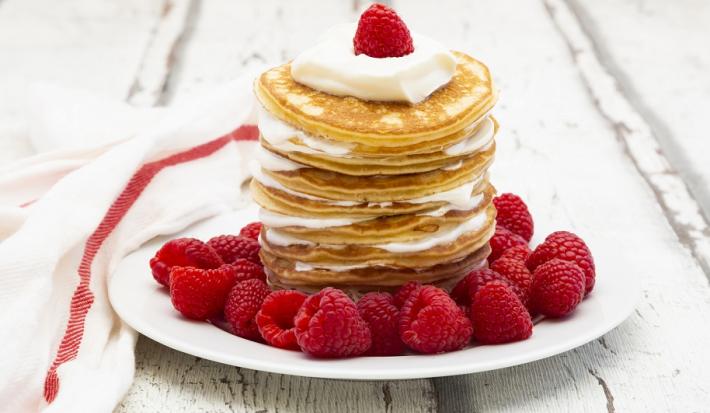 Fotografia em tons de branco em uma bancada de madeira de cor branca. Ao centro, um prato branco contendo as pancakes com frutas espalhadas ao redor. Ao lado, um pano branco espalhado.