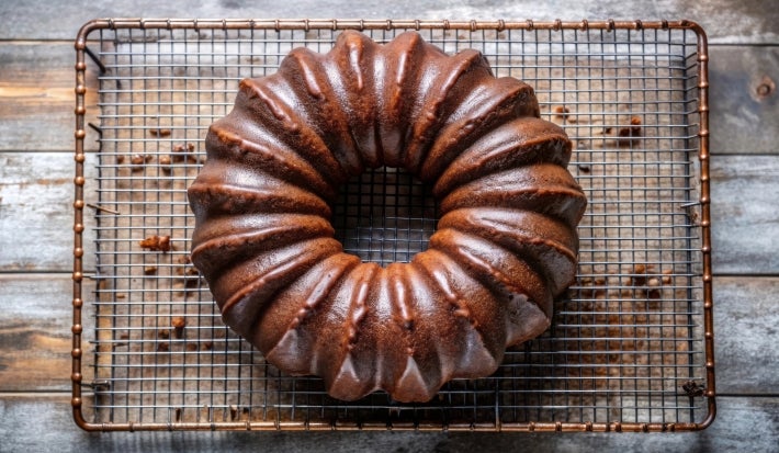 Fotografia de um bolo de chocolate em uma grade de resfriamento, com cor marrom escura e superfície brilhante. O bolo tem um padrão flutuante e está sobre uma superfície de madeira rústica.