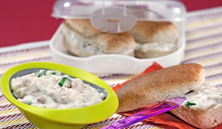 Fotografia em tons de verde, branco e roxo, abaixo potinho com patê cremoso, faca roxa sobre pão aberto ao meio com patê sobre folha de cor salmão, acima recipiente branco com lanches, tudo sobre toalha multicolorida e fundo lilás.