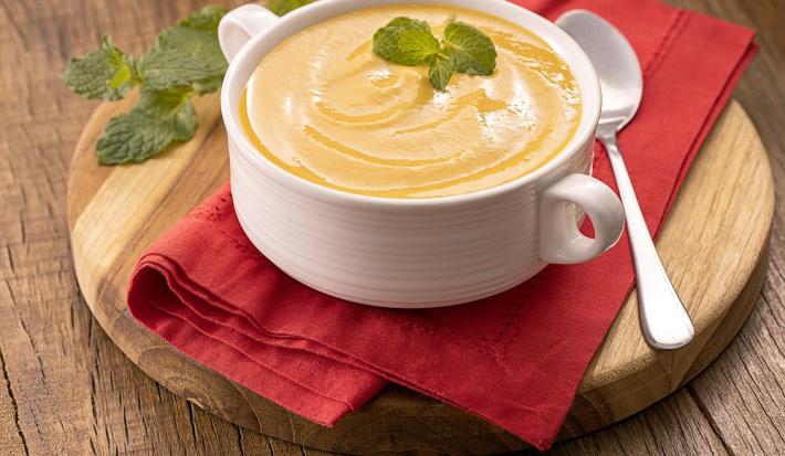 Foto da receita de creme de abóbora com hortelã servida em uma panela de porcelana branca sobre uma tábua de madeira redonda com um pano vermelho entre elas. Para decoração, uma colher prateada ao lado e folhas de hortelã ao fundo.