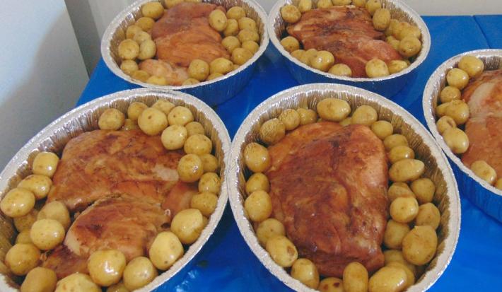Foto da receita de Pernil Assado com Batatas Coradas. Observa-se 5 travessas de alumínio com o pernil dentro bem dourado e muitas batatinhas ao redor.