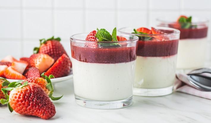 Fotografia em tons de branco em uma bancada de madeira de cor branca. Ao centro, um recipiente contendo a mousse. Ao lado, há alguns morangos espalhados e algumas colheres de inox.