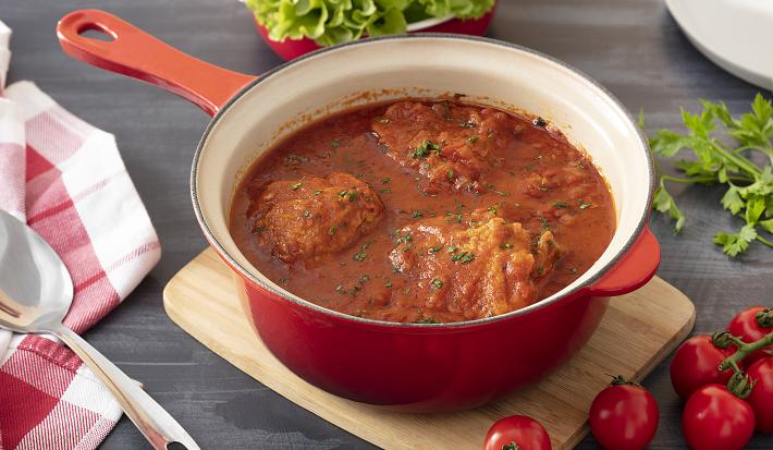 Foto em tons de vermelho da receita de frango com molho de tomate caseiro servida em uma panela vermelha sobre uma tábua de madeira retangular. Ao lado, há tomates e alfaces de decoração, além de um paninho quadriculado branco e vermelho