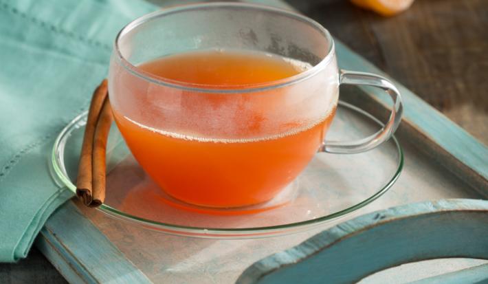 fotografia em tons de laranja, azul e cinza de uma bancada cinza vista de frente. Contém uma bandeja de madeira azul com um prato redondo transparente ao lado uma canela em pau e por cima uma xicara transparente com o chá. Ao lado um pano azul.