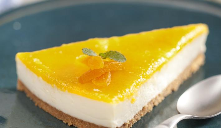 Fotografia em tons de amarelo em uma bancada de madeira com uma toalha cinza, um prato azul redondo raso com uma fatia da torta com cobertura de damasco.