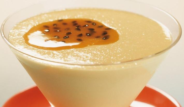 Fotografia em tons de amarelo e vermelho de uma bancada azul e branca com um pratinho vermelho e sobre ele uma taça com o creme de maracujá decorado com polpa e sementes de maracujá.