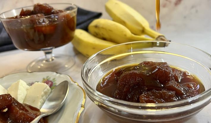 Foto da receita de compota de banana servida uma parte em um bowl de vidro e outra em uma taça baixa atrás. Ao lado, há um prato de porcelana com queijo branco e compota de banana e uma colher prateada. Ao fundo um cacho de bananas e um pano preto.