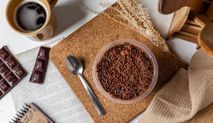 Fotografia em tons de madeira com um pote de plástico ao centro. Dentro do pote existe uma receita vista de cima de um bolo de pole de cenoura com cobertura de brigadeiro e granulados