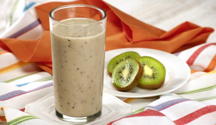 Fotografia em tons de laranja em uma bancada de madeira com uma toalha branca como listras coloridas, um pano laranja, um prato branco redondo pequeno com fatias de kiwi e um copo de vidro alto com o smoothie de iogurte com kiwi e banana.