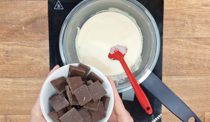 Duas mãos adicionam tabletes de chocolate ao creme de leite cozinhando em banho maria sobre fogão portátil preto.