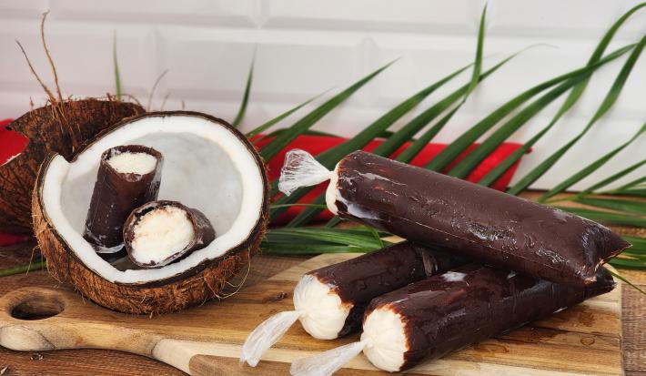 Fotografia de geladinhos em sacos plásticos com cobertura de chocolate e creme de coco branco por dentro. Ao lado cocos decorativos.