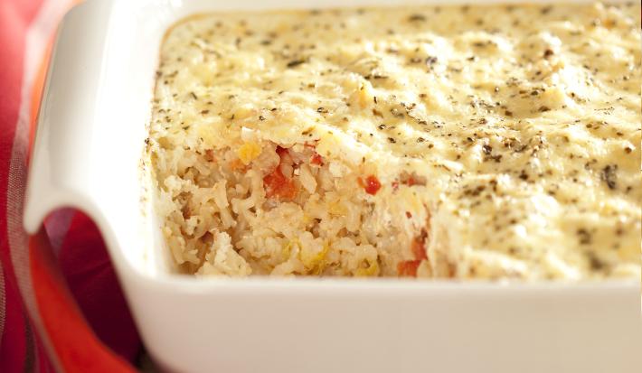 fotografia em tons de marrom, branco e vermelho de uma bancada marrom vista de frente, contém um recipiente quadrado branco com arroz e queijo por cima. Ao lado um pano vermelho com listras roxas.