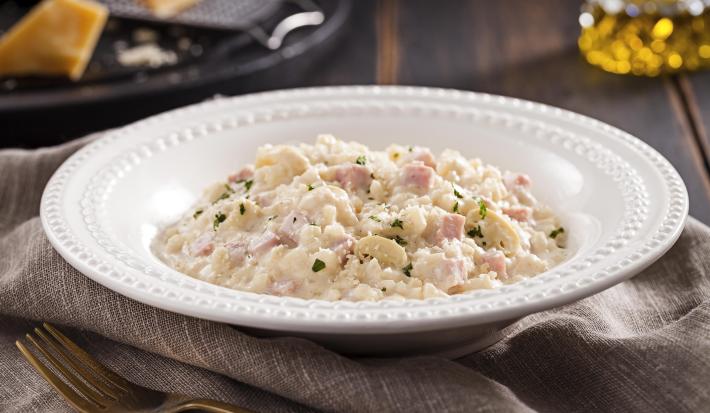 Foto em tons de cinza e branco de uma bancada de madeira escura com uma pano cinza dobrado, um garfo dourado envelhecido e uma prato redondo branco com o risoto dentro. Ao fundo, um prato preto e uma jarrinha de azeite.