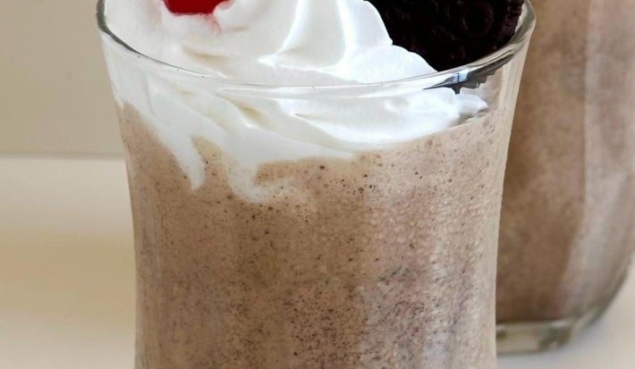 Fotografia em tons claros de dois copos de vidro com milkshake de negresco, decorados com chantilly, cereja e um biscoito Negresco inteiro.