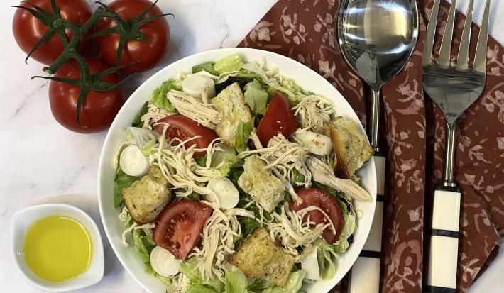 Foto vista de cima da receita de Salada de Frango servida em um recipiente grande, com um recipiente menor com Molho de Mostarda. Tudo está sobre uma bancada decorada com um tecido vermelho, um par de talheres e alguns tomates