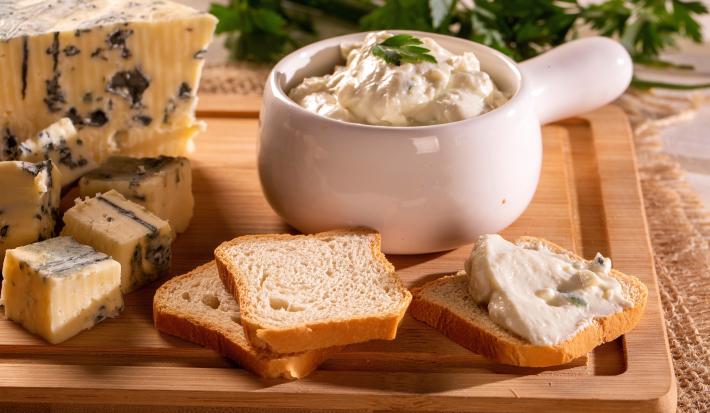 Fotografia de um recipiente pequeno de vidro branco com patê de gorgonzola de damasco, ao lado de uma peça e alguns pedaços de gorgonzola e algumas fatias de torrada, que estão sobre uma tábua de madeira.