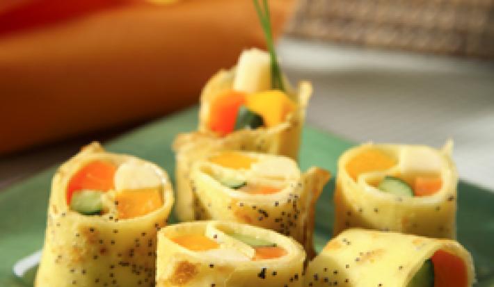 Fotografia em tons de laranja, amarelo e verde, com pedaços de panqueca recheadas com legumes sobre peça de vidro em tom verde, ao lado guardanapo laranja, ao fundo guardanapos de cores variadas, tudo sobre bancada em tom cinza.